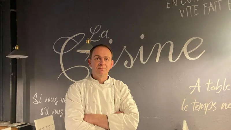 Chef de braços cruzados em frente à parede cinza com a inscrição "La Cuisine" no restaurante em Punta Arenas.