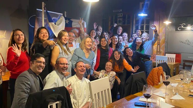 Grupo animado posando no restaurante La Cuisine em Punta Arenas, com bandeira chilena ao fundo e taças de vinho na mesa.