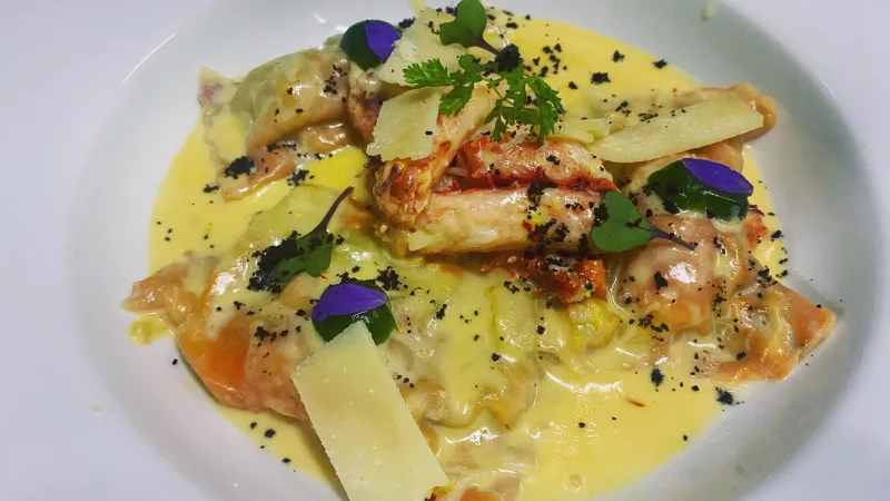 Prato refinado com massa recheada, frutos do mar, molho cremoso amarelo, lascas de queijo e flores comestíveis roxas