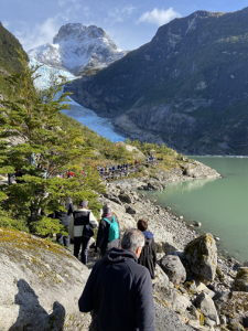 Grupo de visitantes caminando por sendero rocoso hacia el glaciar Serrano, con lago glaciar turquesa y montañas nevadas al