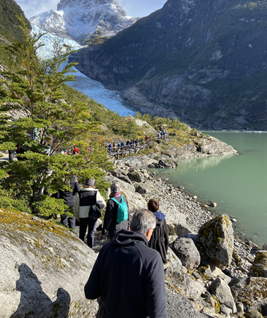 Grupo de visitantes caminando por sendero rocoso hacia el glaciar Serrano, con lago glaciar turquesa y montañas nevadas al