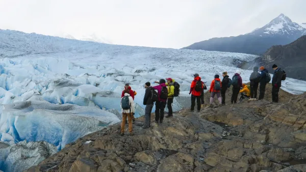 A group of hikers with backpacks standing on rocky terrain at the edge of Grey Glacier, with blue ice and mountains behind