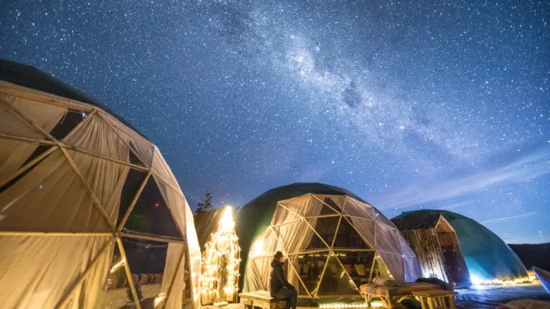Geodesic dome glamping tents lit with fairy lights at night under a stunning Milky Way starry sky, person sitting outside