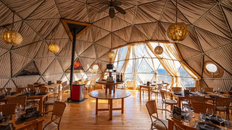 Interior of EcoCamp Patagonia geodesic dome restaurant with wicker chairs, wooden tables, warm globe lights, and panoramic