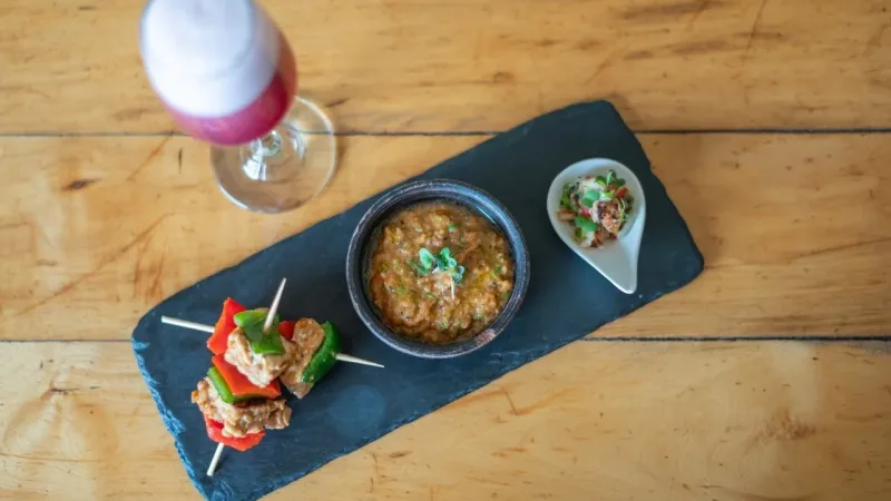 Appetizer slate board with vegetable skewers, a dip bowl, and a small bite, beside a pink foamy cocktail on a wood table