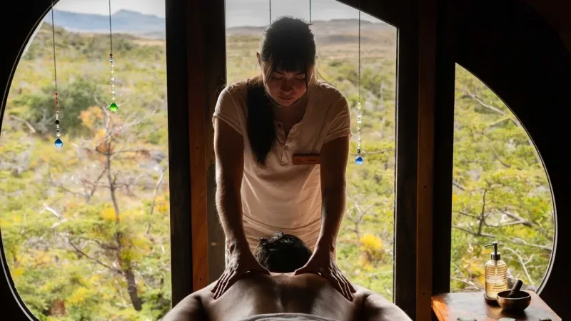 Spa therapist giving a back massage in a room with a large circular window overlooking a lush mountain landscape