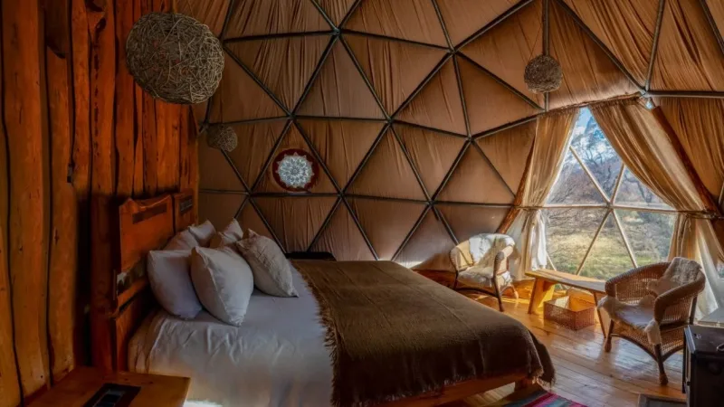 Interior of an EcoCamp Patagonia geodesic dome with a double bed, wicker chairs, colorful rug, and Patagonia landscape view