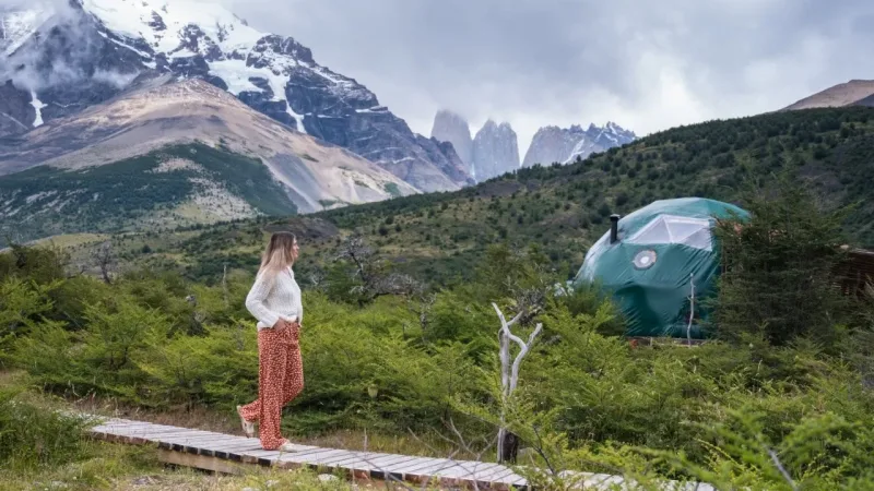 Woman standing on a wooden walkway at EcoCamp Patagonia, with a green dome tent and Torres del Paine peaks behind her.
