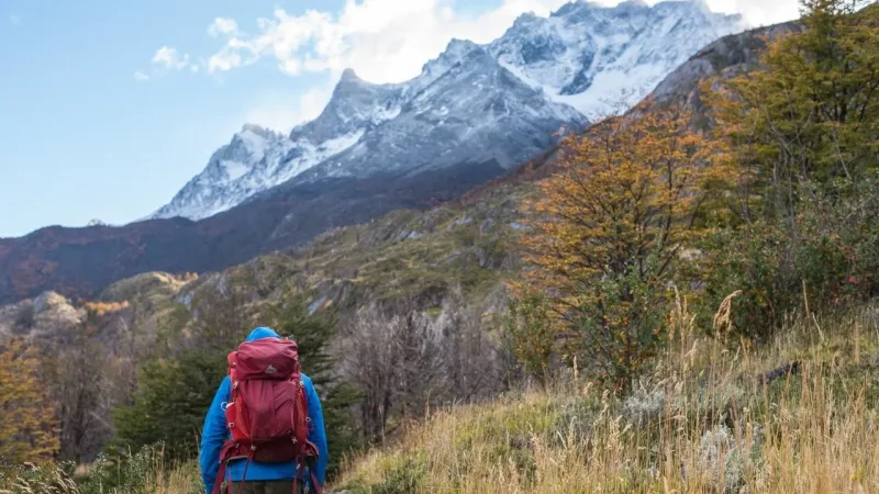 Hiker with red backpack on a Patagonia trail surrounded by autumn trees and snow-capped mountain peaks