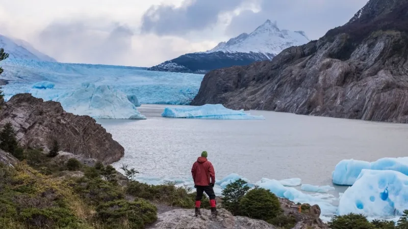 Hiker in red jacket standing on rocks overlooking Grey Glacier with floating icebergs and snowy peaks in Patagonia