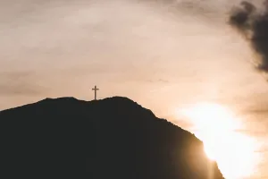 Cruz en la cima del Cabo Froward en silueta, con el sol poniéndose detrás del cerro y cielo nublado.