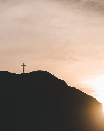 Cruz en la cima del Cabo Froward en silueta, con el sol poniéndose detrás del cerro y cielo nublado.