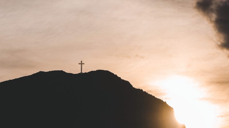Cruz en la cima del Cabo Froward en silueta, con el sol poniéndose detrás del cerro y cielo nublado.