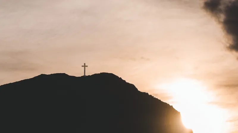 Cruz en la cima del Cabo Froward en silueta, con el sol poniéndose detrás del cerro y cielo nublado.
