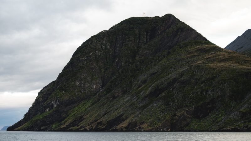 Cabo Froward visto desde el mar, con una cruz en su cima rocosa cubierta de vegetación bajo cielo nublado.