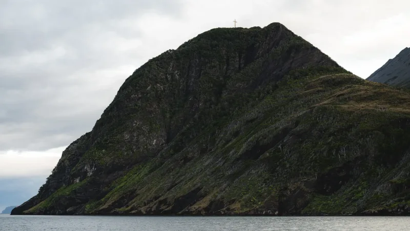 Cabo Froward visto desde el mar, con una cruz en su cima rocosa cubierta de vegetación bajo cielo nublado.