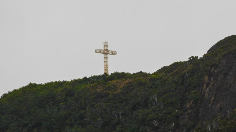 Cruz metálica dorada en la cima del Cabo Froward, rodeada de vegetación patagónica bajo un cielo gris
