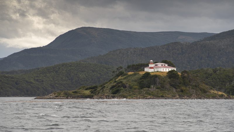 Faro San Isidro sobre un promontorio verde rodeado de mar agitado, con montañas y cielo nublado al fondo.