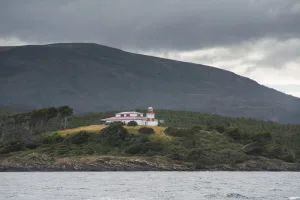 Faro San Isidro de color blanco y rojo sobre una colina costera rodeada de vegetación, con montañas nubladas al fondo.