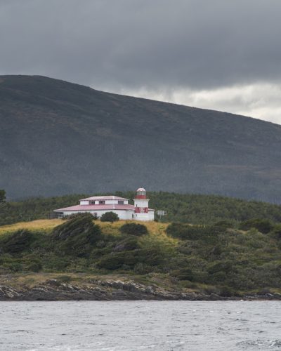 Faro San Isidro de color blanco y rojo sobre una colina costera rodeada de vegetación, con montañas nubladas al fondo.