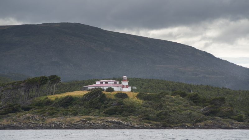 Faro San Isidro de color blanco y rojo sobre una colina costera rodeada de vegetación, con montañas nubladas al fondo.